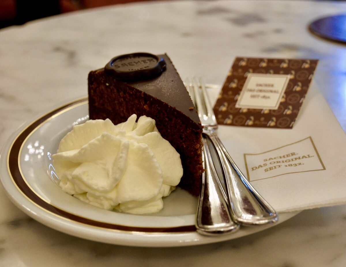 A slice of Sachertorte with whipped cream and the round chocolate seal of Hotel Sacher