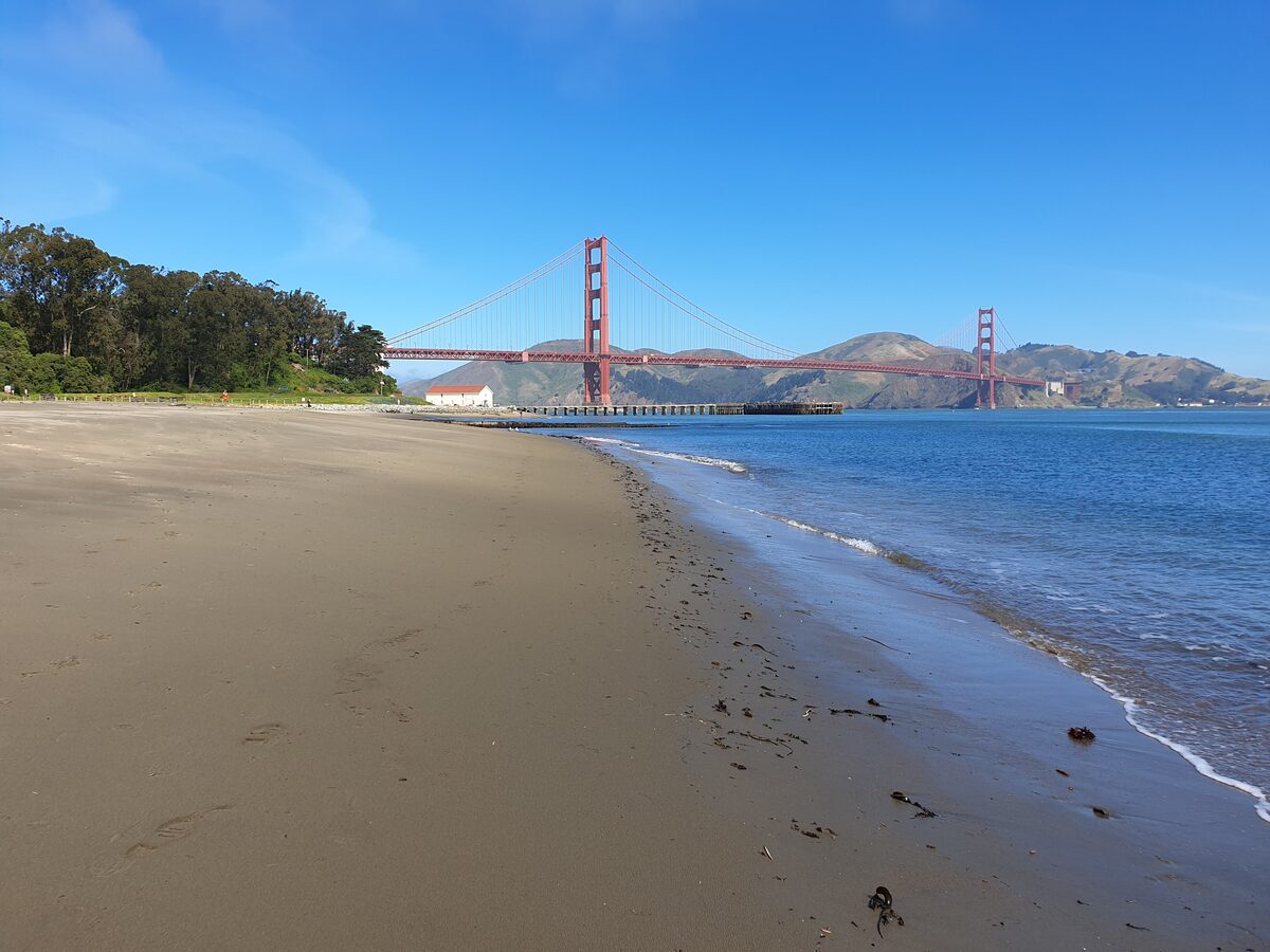 The sandy shore of Crissy Field with the Golden Gate Bridge in the distance under a blue sky