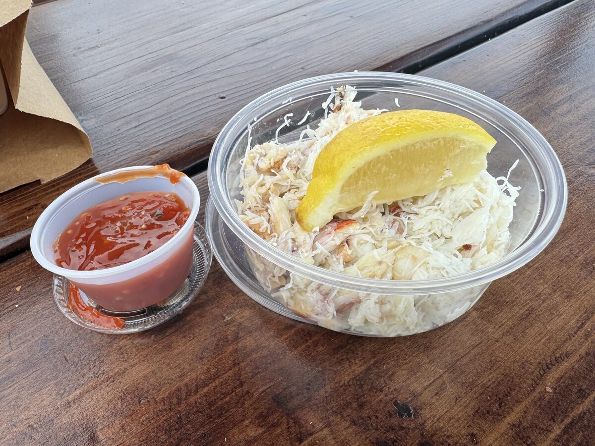 A takeaway cup of fresh crab meat with a lemon wedge and cocktail sauce on a wooden table