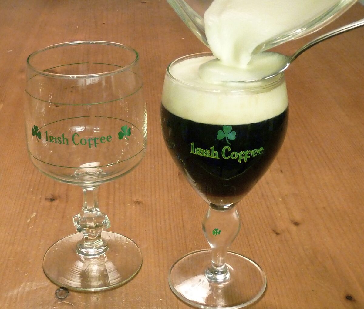 Cream being poured over hot coffee in a stemmed Irish coffee glass