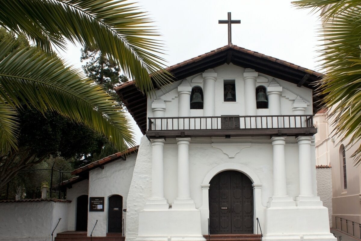 The whitewashed adobe front of Mission Dolores with its columns, balcony and cross