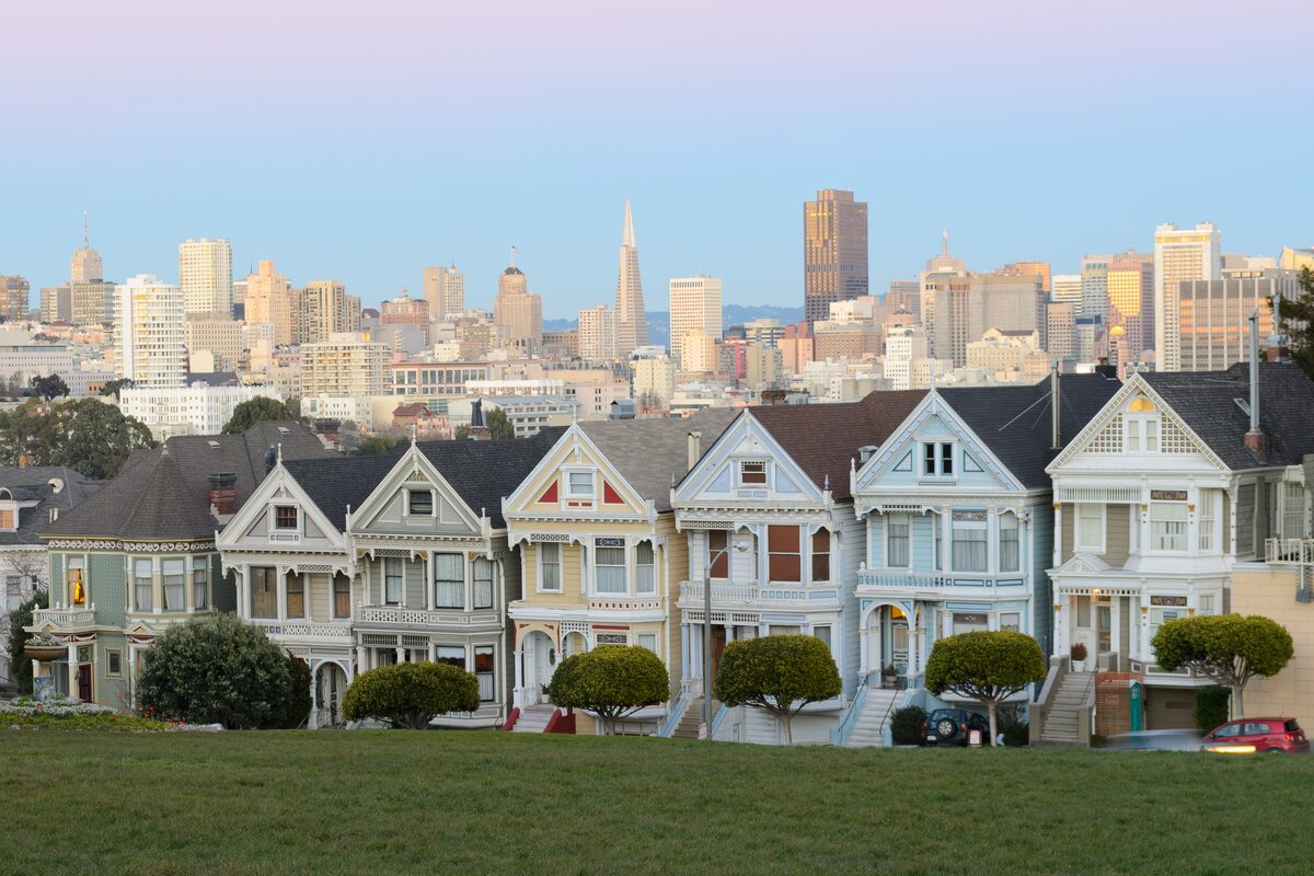 A row of pastel Victorian houses, the Painted Ladies, on the edge of Alamo Square with the downtown skyline behind