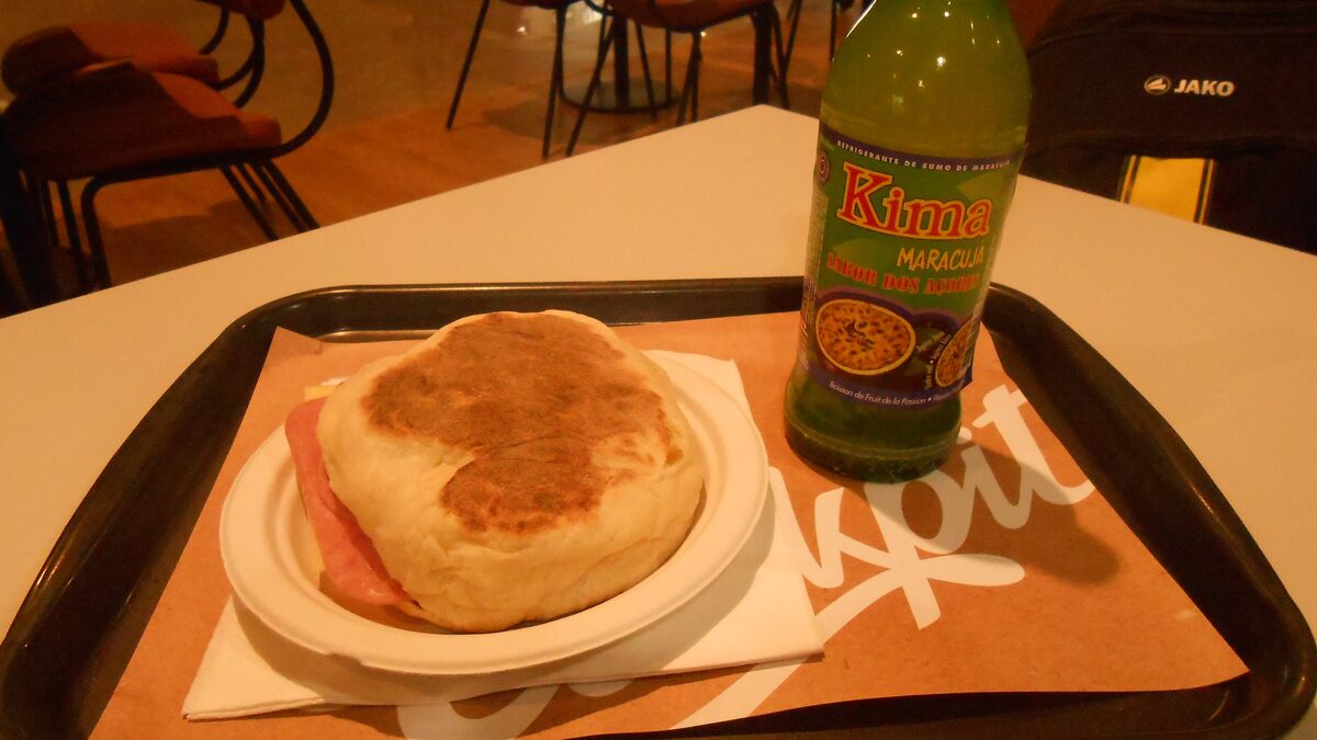 A round bolo lêvedo bun split open with ham on a café tray, next to a bottle of Kima