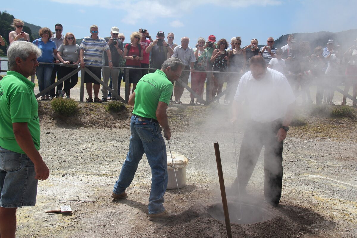 Visitors watch two men lift a cozido pot out of the steaming ground in Furnas