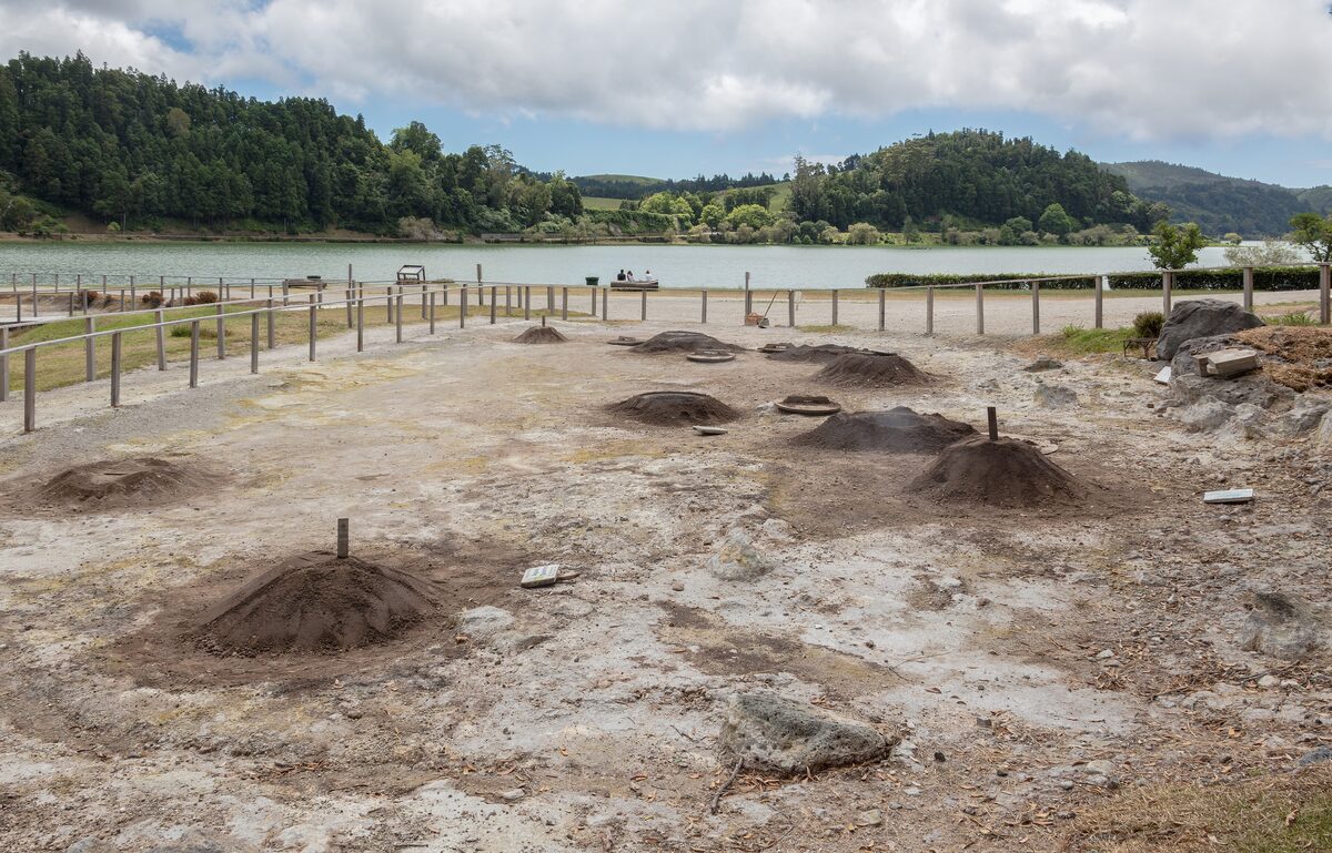 Steam vents and fumarole mounds on the ground beside Lagoa das Furnas, with the lake and its green hills behind