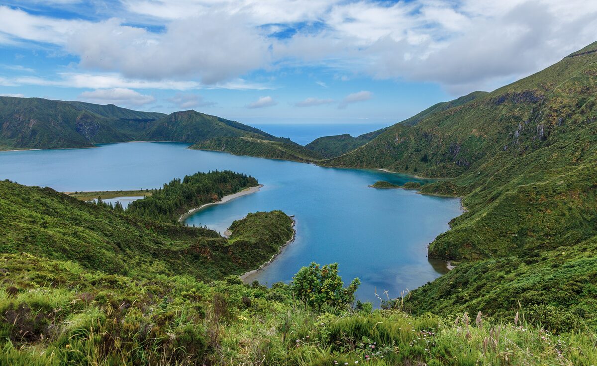 The blue crater lake of Lagoa do Fogo seen from above, with a forested peninsula, a sandy shore and steep green slopes under a bright sky