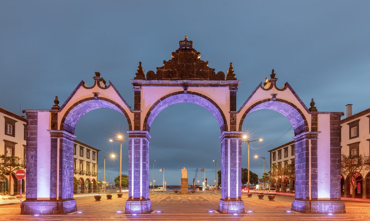 The three lit arches of the Portas da Cidade gate in Ponta Delgada at blue hour, with the street running down toward the sea