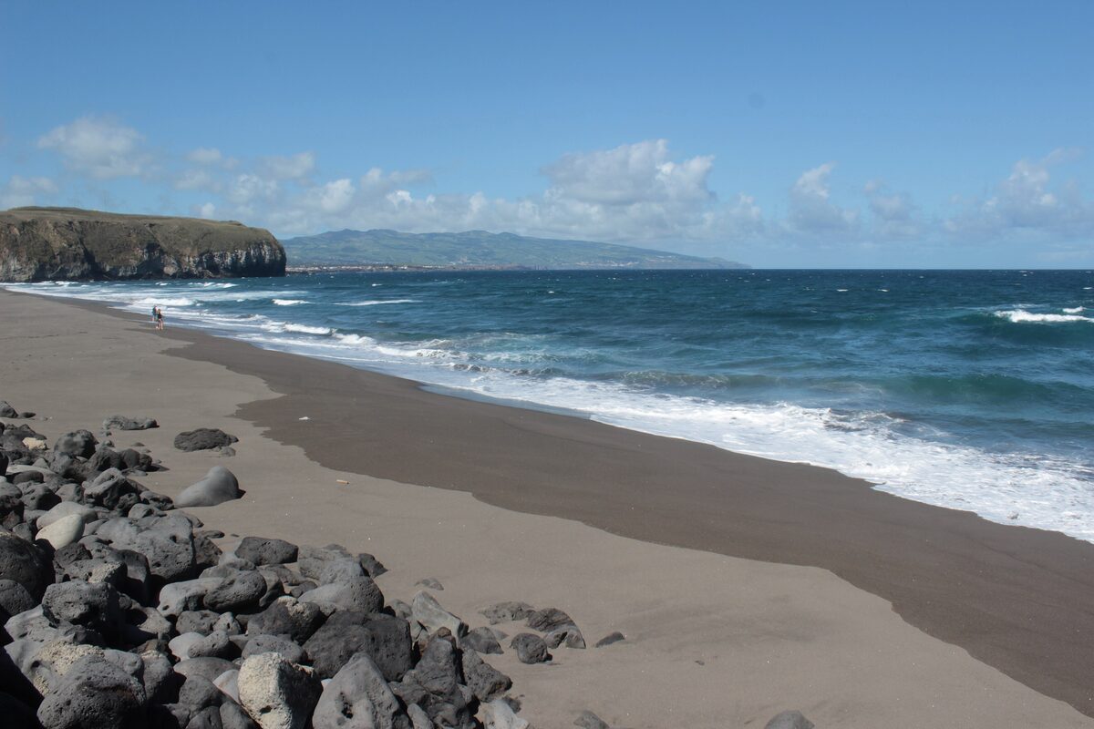 Waves rolling onto the long dark volcanic sand of Santa Bárbara beach, with a cliff headland and the coast beyond