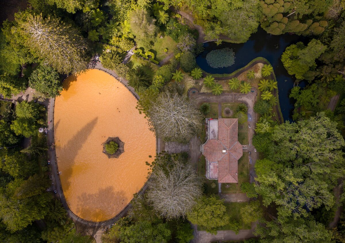 Aerial view of the orange, iron-rich thermal pool in Terra Nostra Park, with its small island, the garden house and dense trees around it