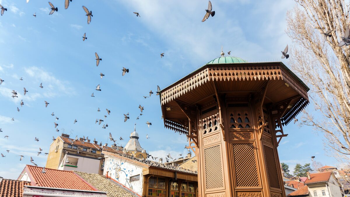 The Sebilj fountain on Baščaršija square with a flock of pigeons in flight and old-town roofs behind it