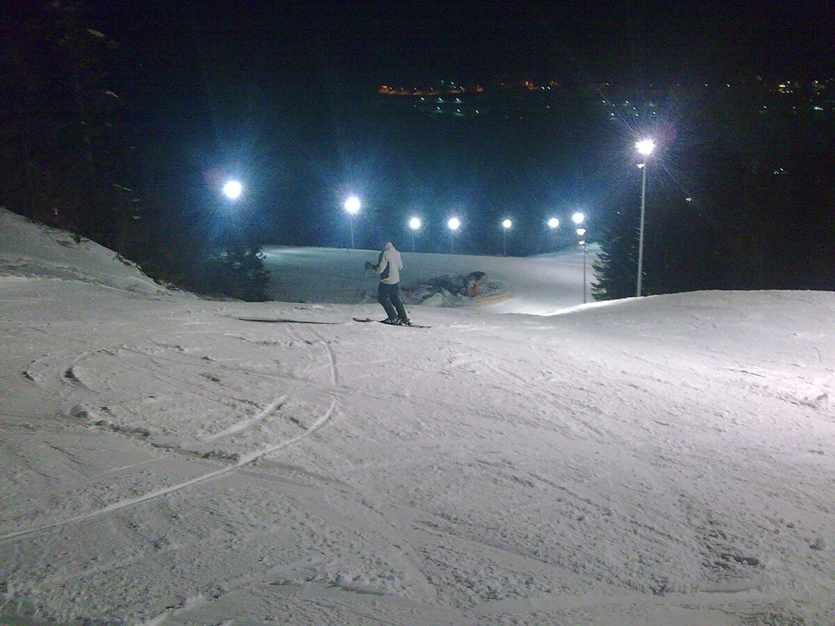 A skier on a floodlit, groomed piste at night on Bjelašnica, with lamp posts lining the run