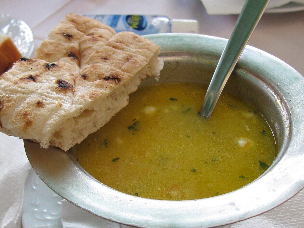 A pewter bowl of creamy yellow begova čorba with a spoon and a piece of flatbread on the rim