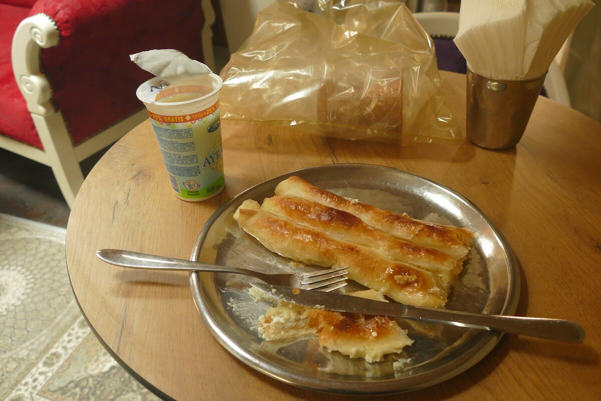 Cheese burek on a metal plate with a fork and a cup of drinking yoghurt on a table beside it