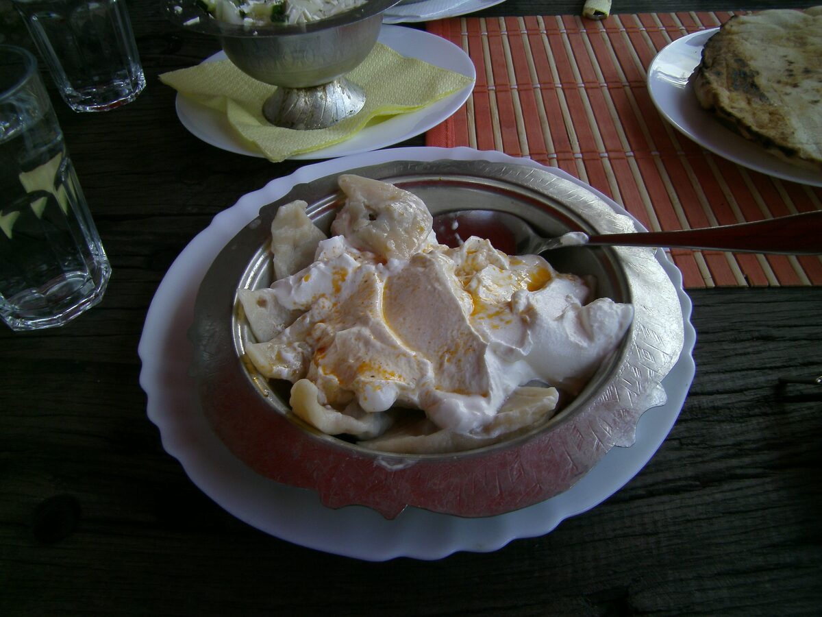 A metal bowl of klepe, Bosnian meat dumplings, covered in white garlic yogurt with a drizzle of paprika butter