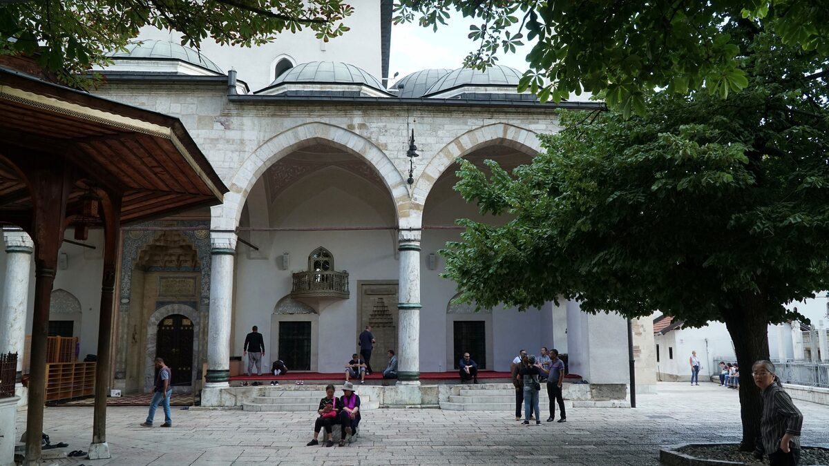 The arcaded courtyard of Gazi Husrev-beg Mosque, with white arches, a tree and people sitting on the stone floor