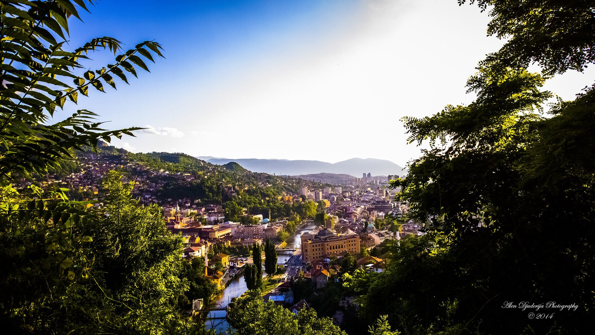 The old town of Sarajevo seen through trees, red roofs stepping down the Miljacka valley toward the hills