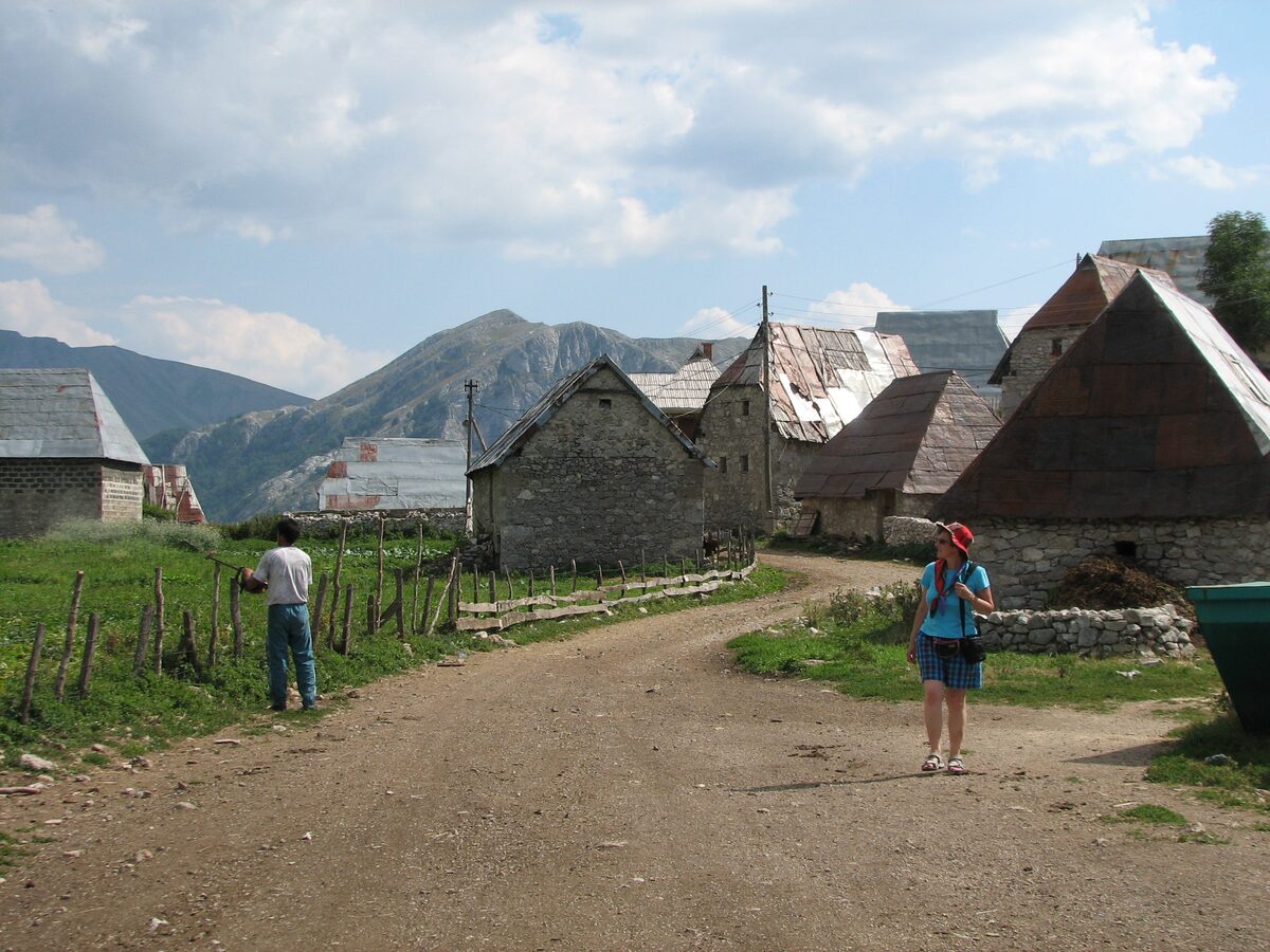 Stone houses and a dirt track in the village of Lukomir, with a hiker on the path and mountains behind