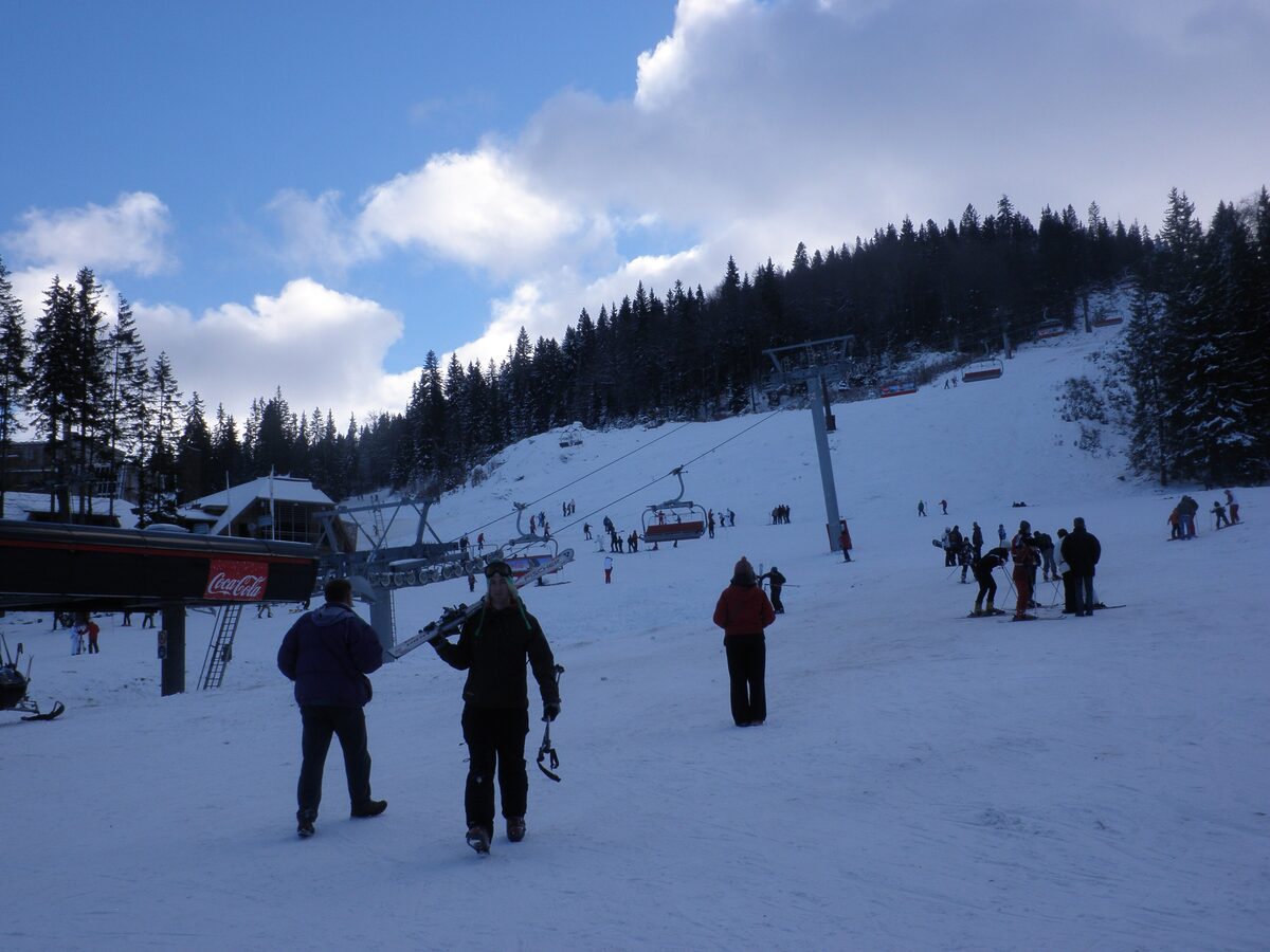 Skiers walking away from the chairlift station at the foot of the Poljice slopes on Jahorina, with spruce forest above the piste