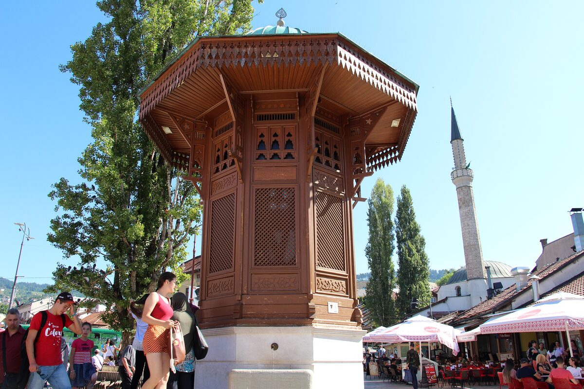 The wooden Sebilj fountain in Baščaršija with a minaret and crowds on the square behind it