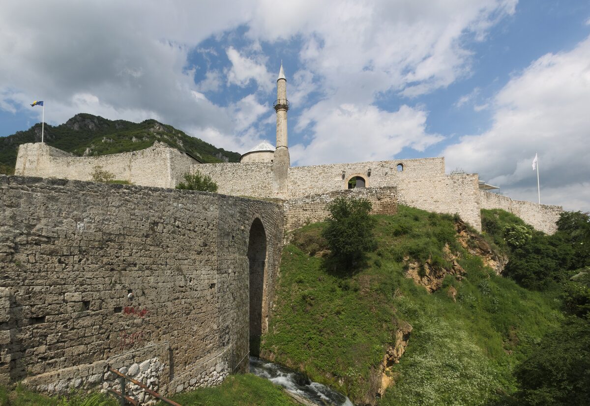 The stone walls of the Travnik fortress on a hill, with a minaret rising behind the ramparts under cloud