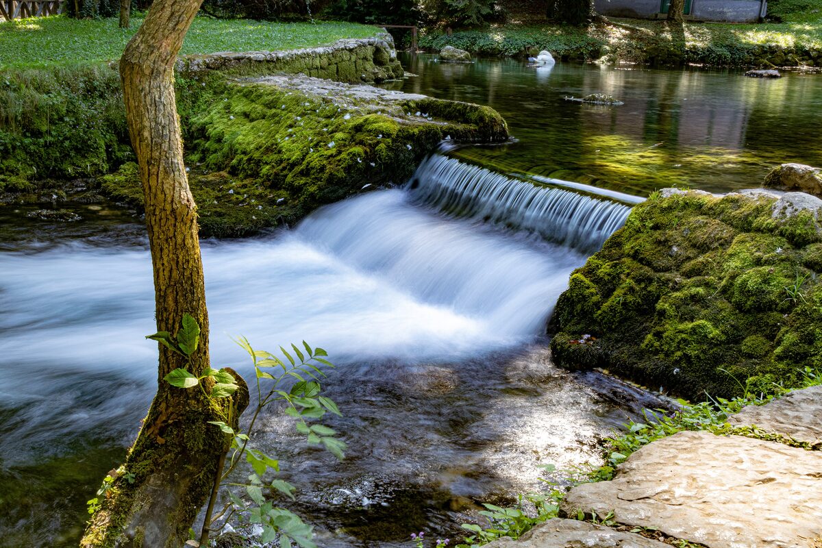 A weir on a stream in the Vrelo Bosne park, with moss-covered rocks and a bare tree on the bank