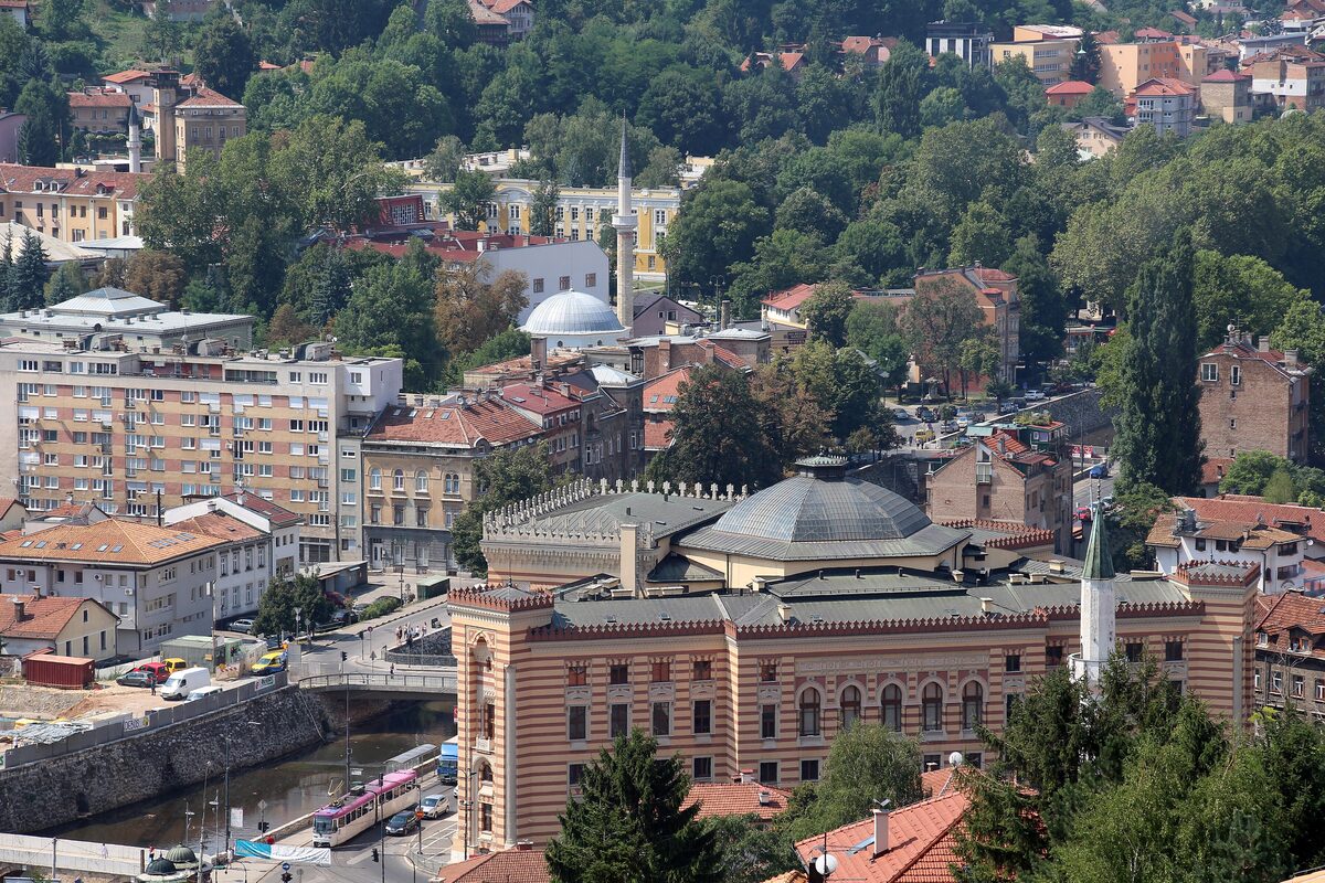 The view from the Yellow Fortress hill, not the fortress itself: the striped City Hall beside the Miljacka, with red roofs and minarets climbing the hill behind