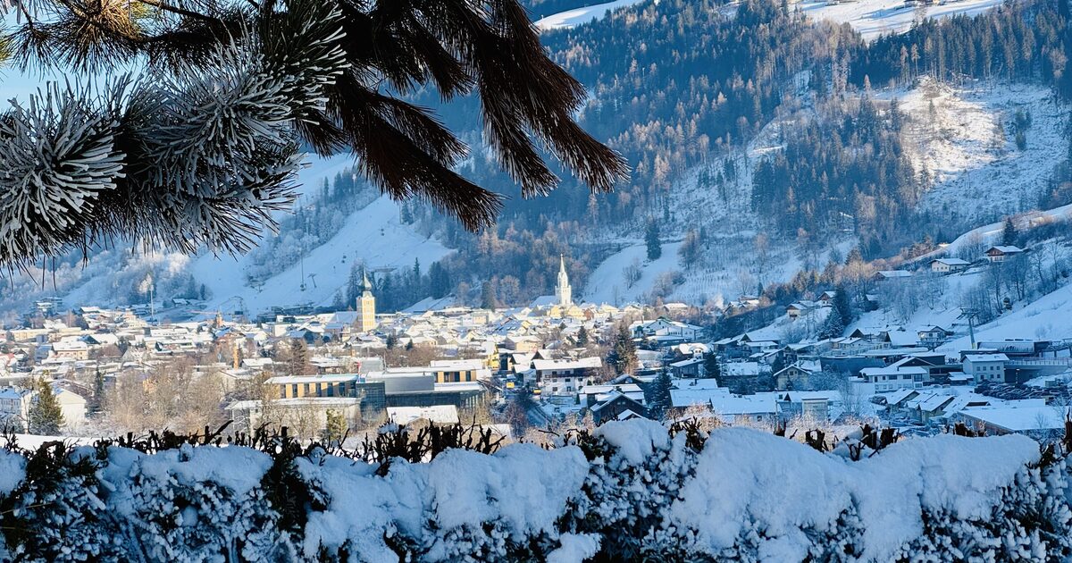 Schladming in winter sun, with snowy roofs, the church towers and the ski slopes above the town, seen through a frosted pine branch