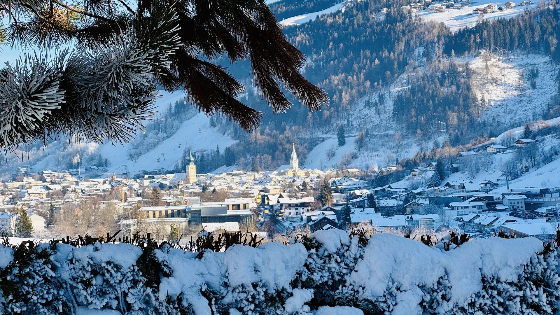 Schladming in winter sun, with snowy roofs, the church towers and the ski slopes above the town, seen through a frosted pine branch