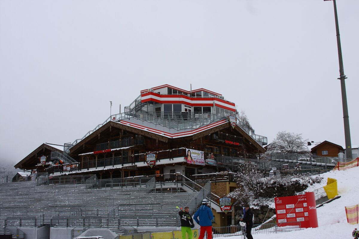 The Hohenhaus Tenne under a grey winter sky, a timber and glass building with a red and white top floor above the empty grandstands of the Planai finish stadium