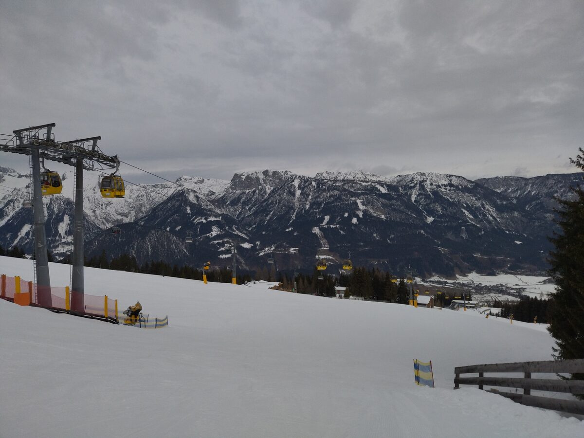Yellow gondola cabins on a lift line above a wide, empty snowy piste on the Hauser Kaibling, with snowy mountains beyond under a grey sky