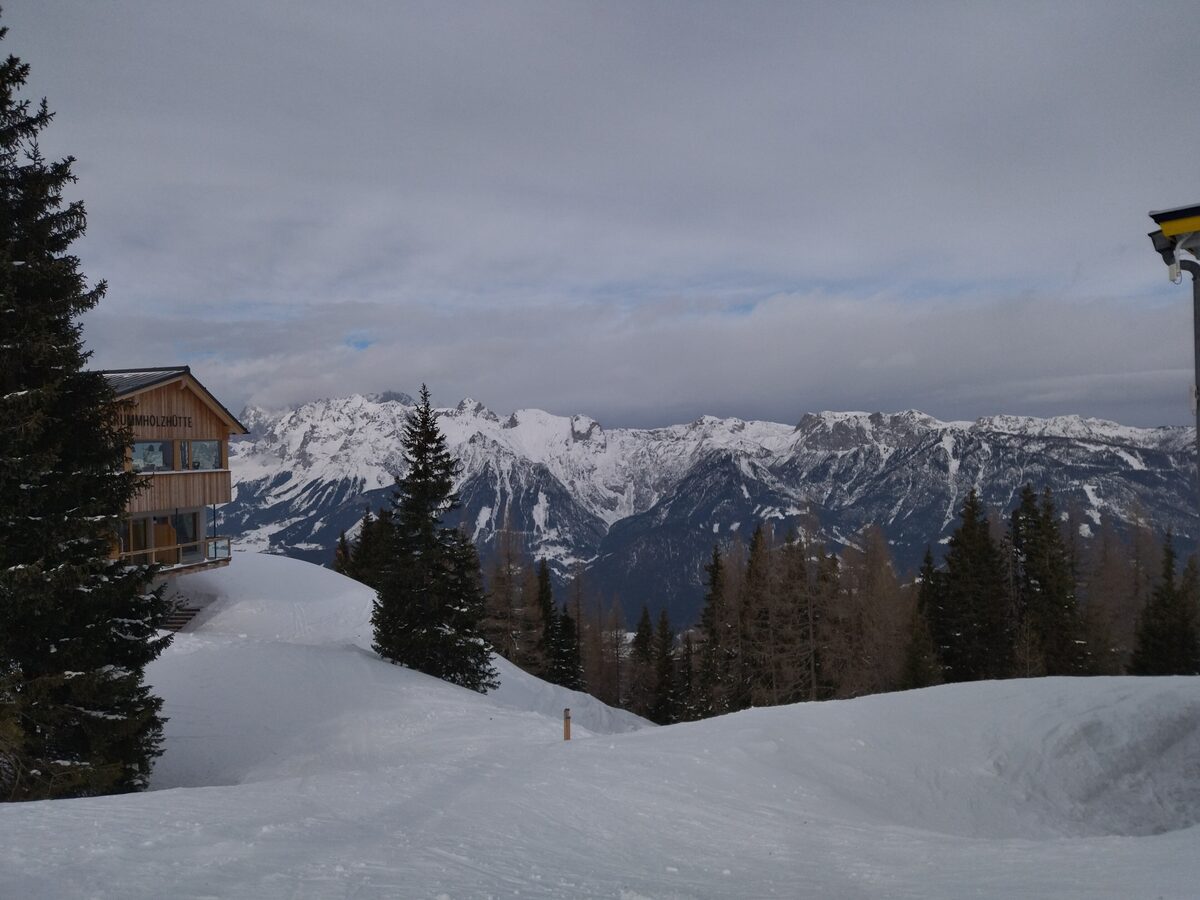 The Krummholzhütte mountain hut on the Hauser Kaibling above a snowy slope, with conifers and the snow-covered Dachstein range under a cloudy sky