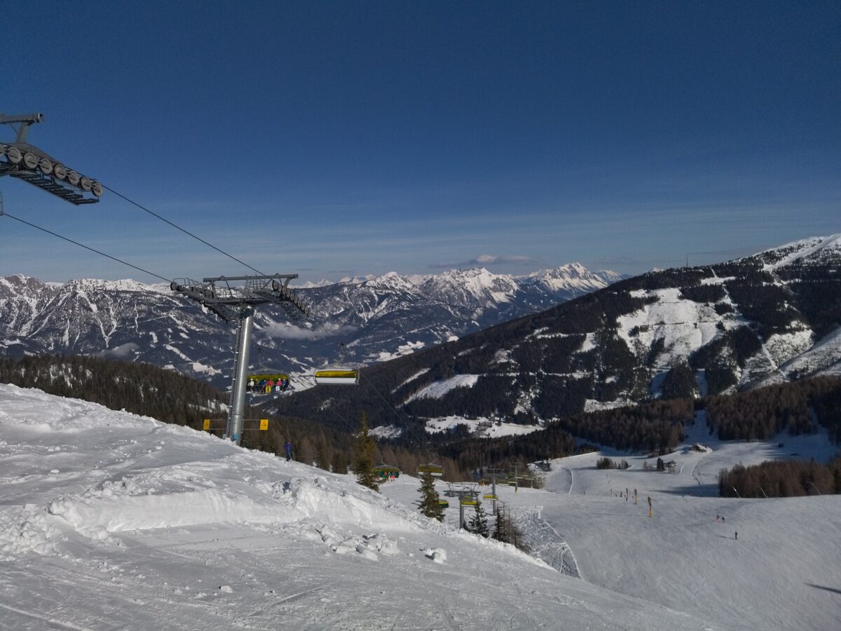 A chairlift above snowy Planai pistes, with skiers on the slopes and snowy mountains beyond