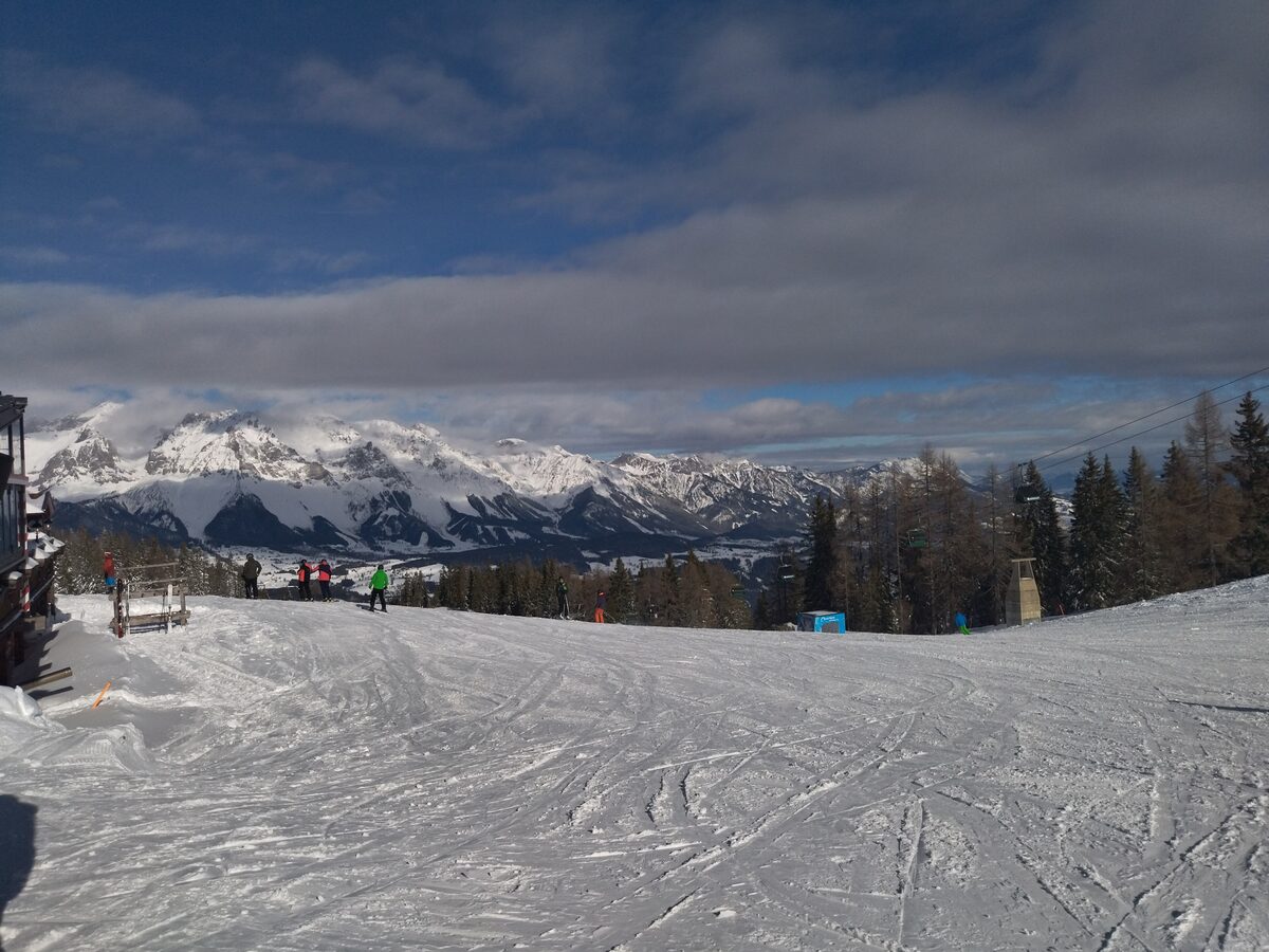 A wide, much-skied piste on the Reiteralm with a few skiers at a hut on the left and the snowy Dachstein range in the distance