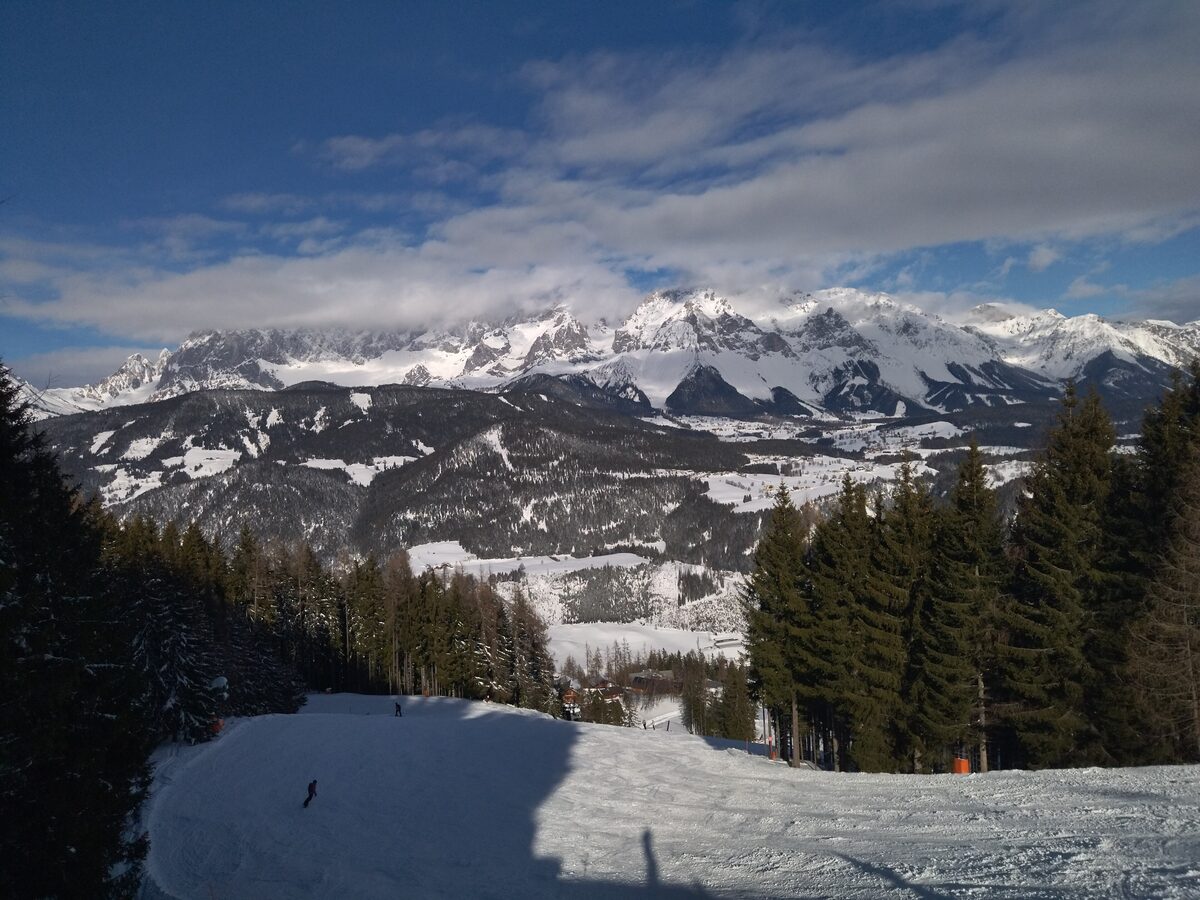 A groomed piste on the Reiteralm in winter light, with spruces on the right and a snow-covered mountain range under a blue sky beyond