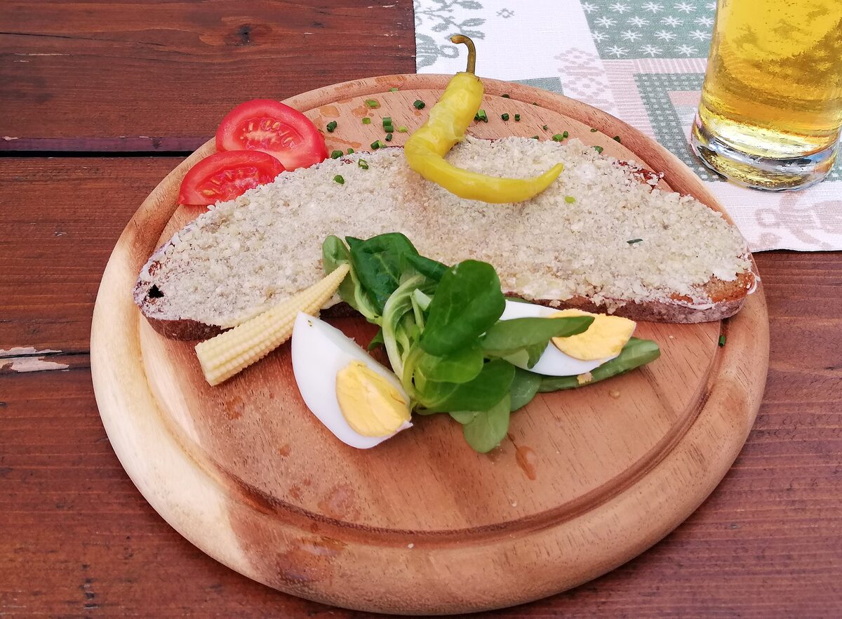 A slice of bread thickly spread with Styrian cheese, served on a wooden board with tomato, a chili pepper, egg, mini corn and salad, and a glass of beer