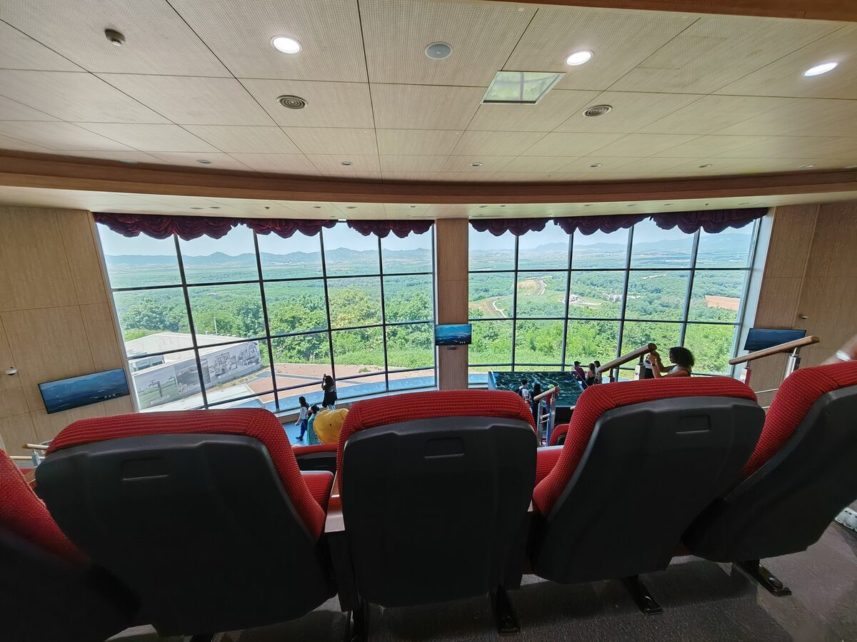 Red seats facing the tall windows of the indoor viewing hall at Dora Observatory, with the green hills of the DMZ outside