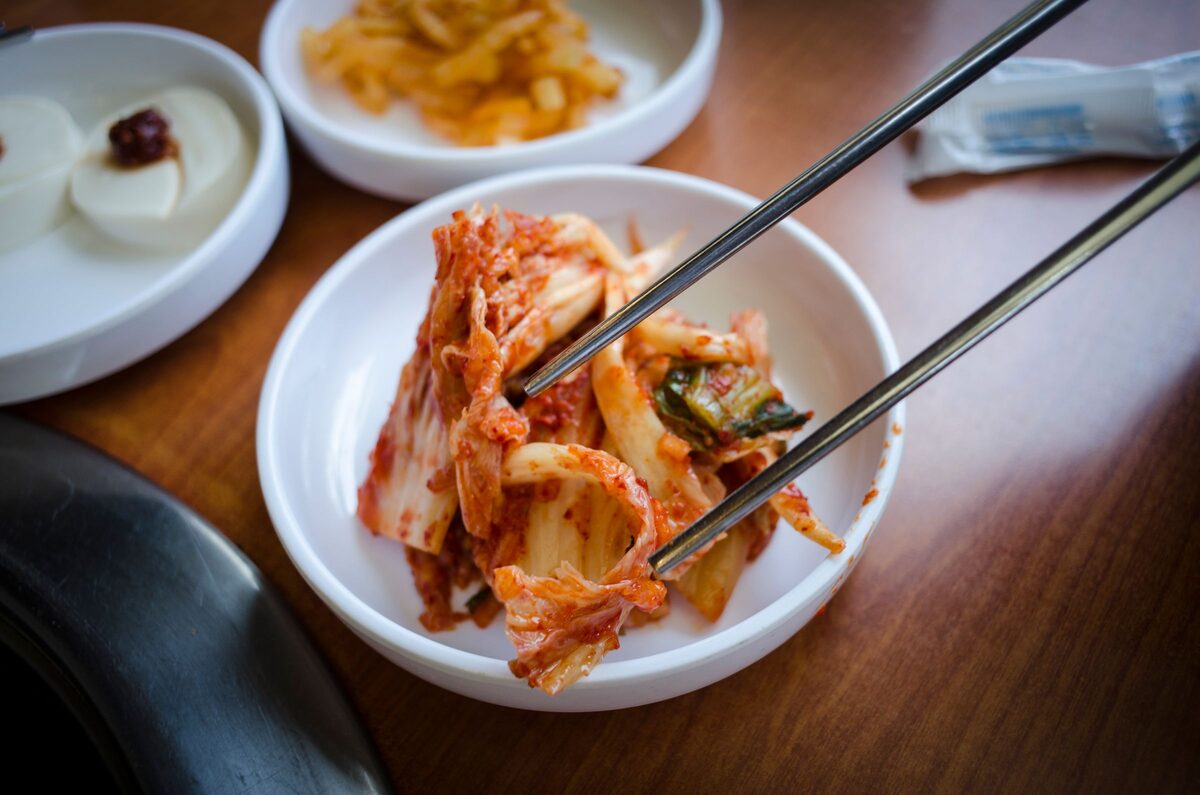 Red napa cabbage kimchi in a white dish, lifted with chopsticks, among other side dishes on a wooden table