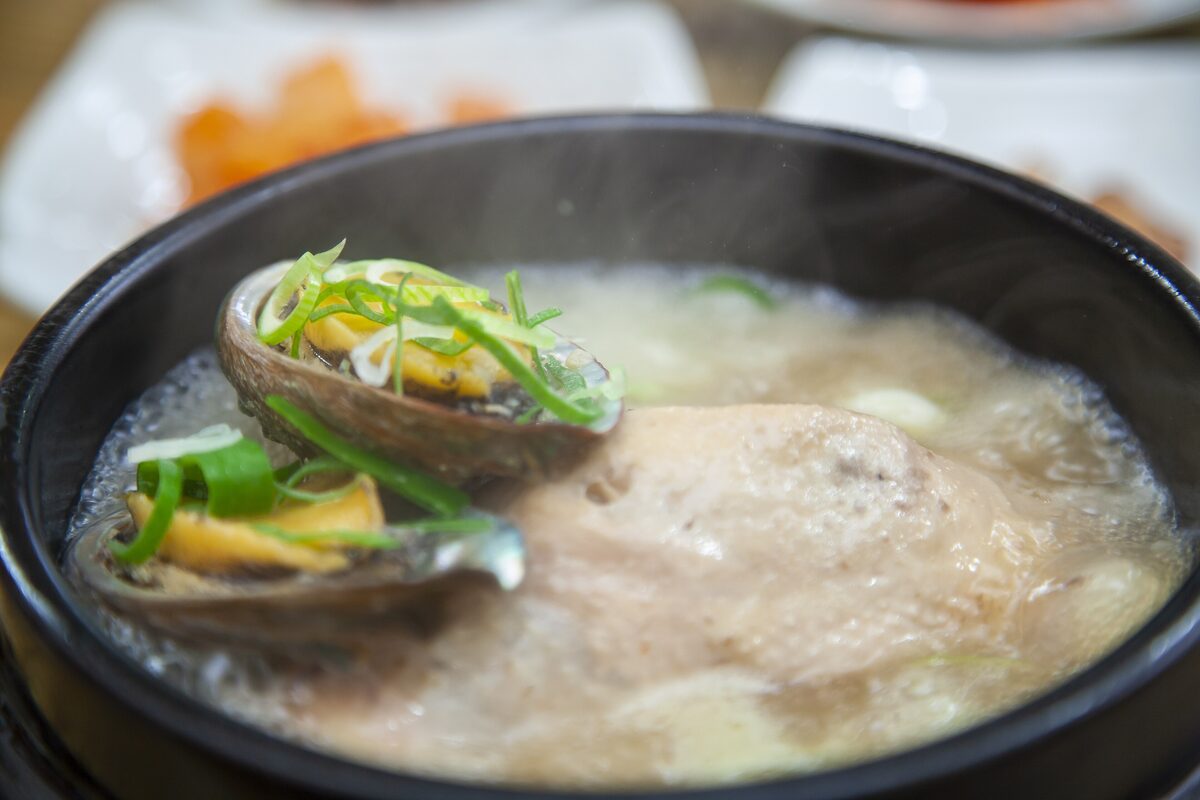A black clay pot of samgyetang, a whole chicken in clear broth with abalone on top