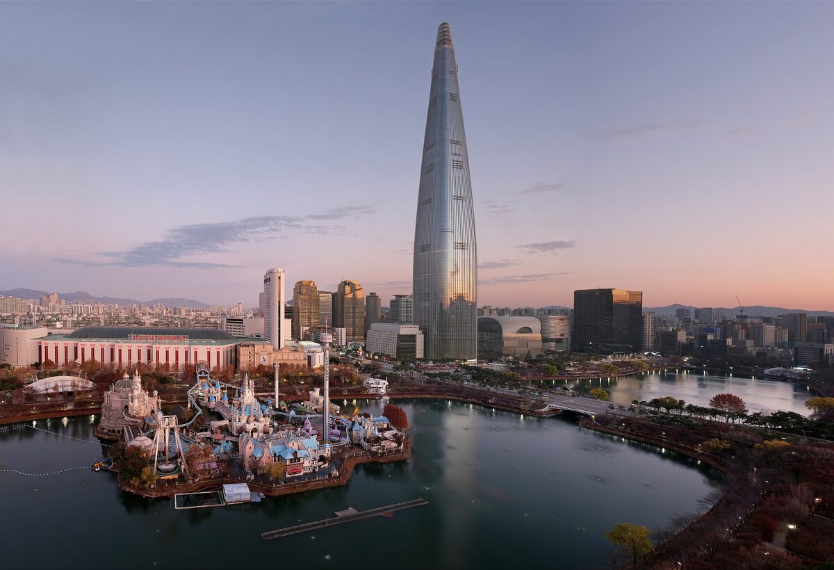 Lotte World Tower rising above Seokchon Lake at dusk, with the lake and the amusement park in the foreground