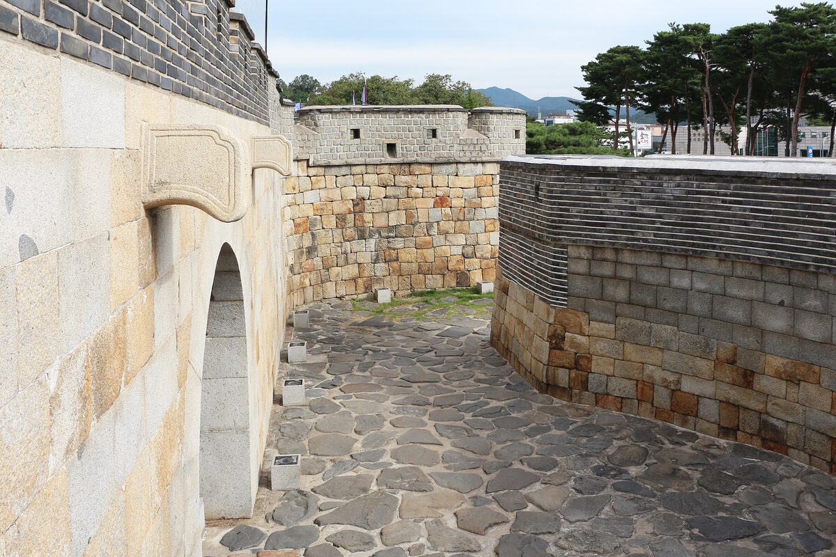 A stone arch gate and wall of Hwaseong Fortress in Suwon, with pine trees beyond