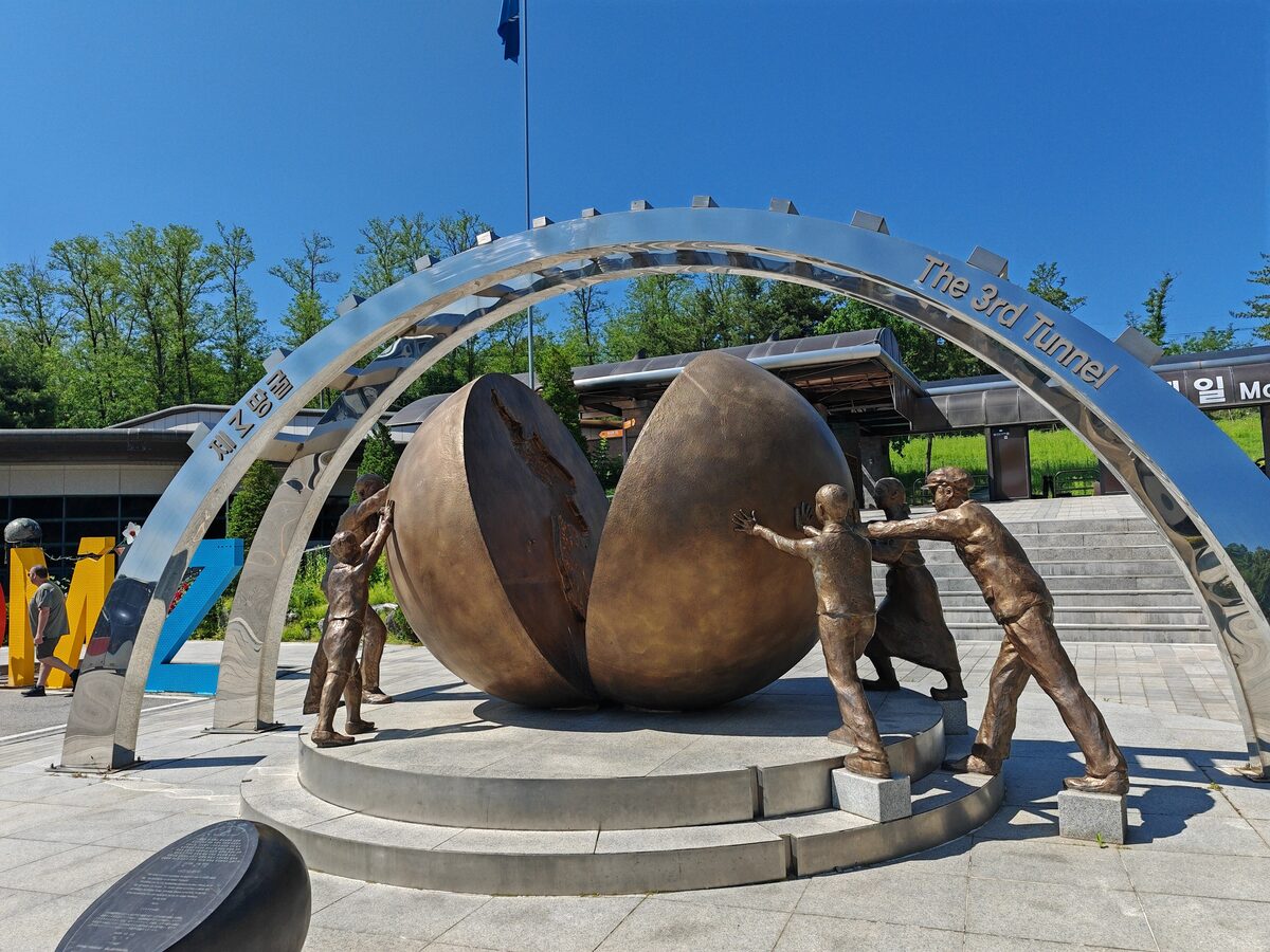 Bronze figures pushing apart a split globe under a steel arch marked The 3rd Tunnel, at the entrance area of the tunnel site