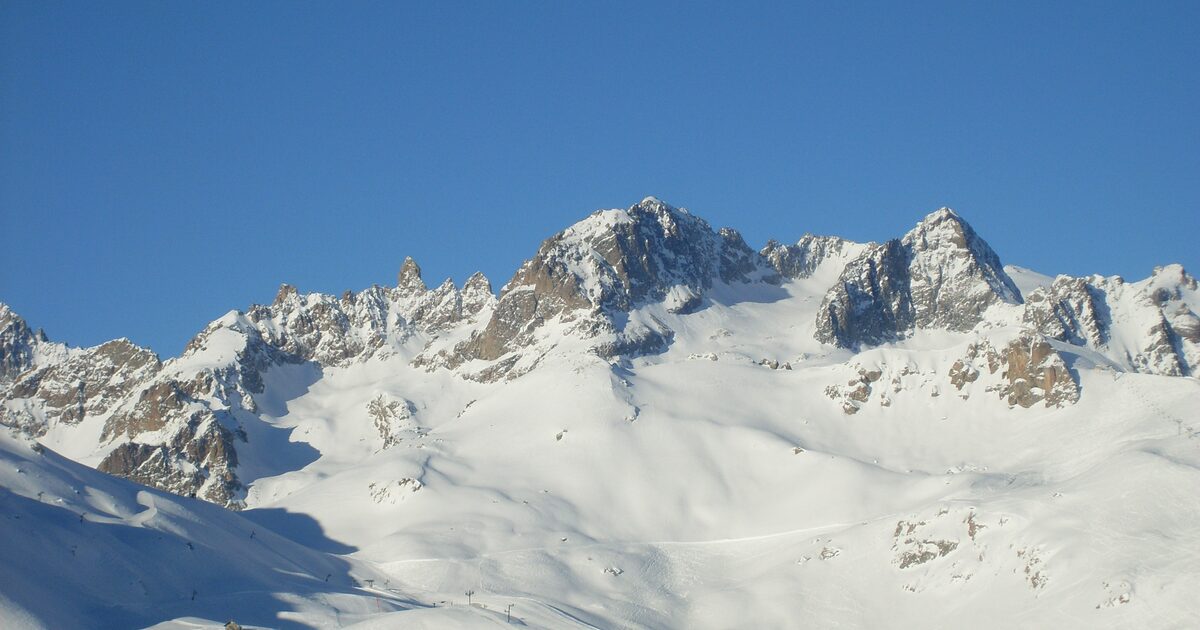 Snowy peaks and rock faces above the Serre Chevalier ski area under a clear blue winter sky, with groomed pistes below
