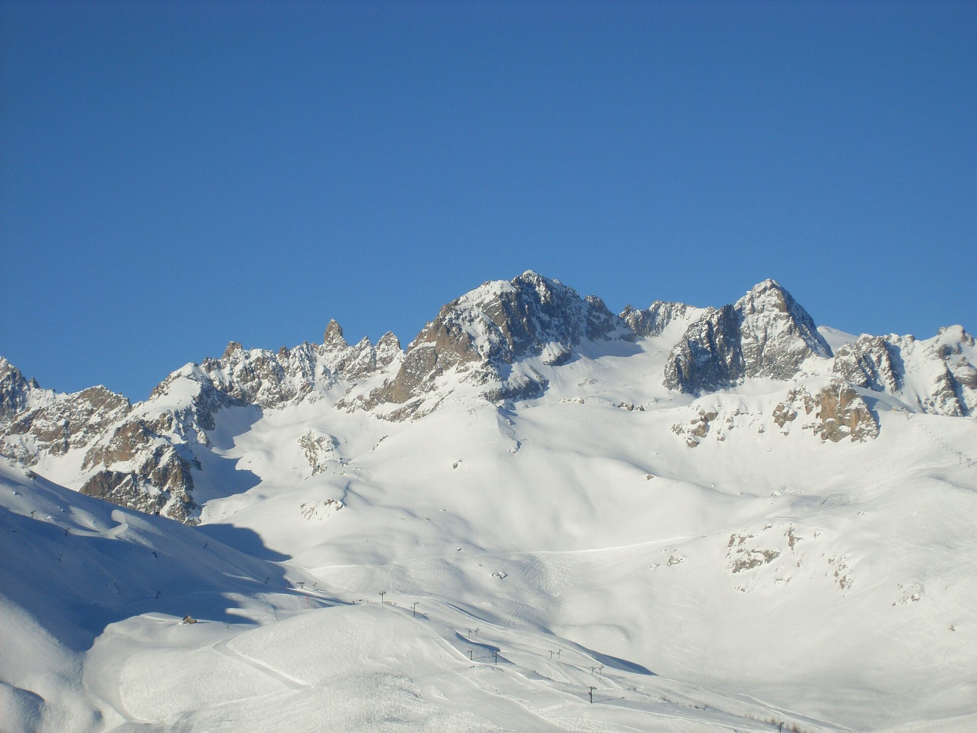 Snowy peaks and rock faces above the Serre Chevalier ski area under a clear blue winter sky, with groomed pistes below