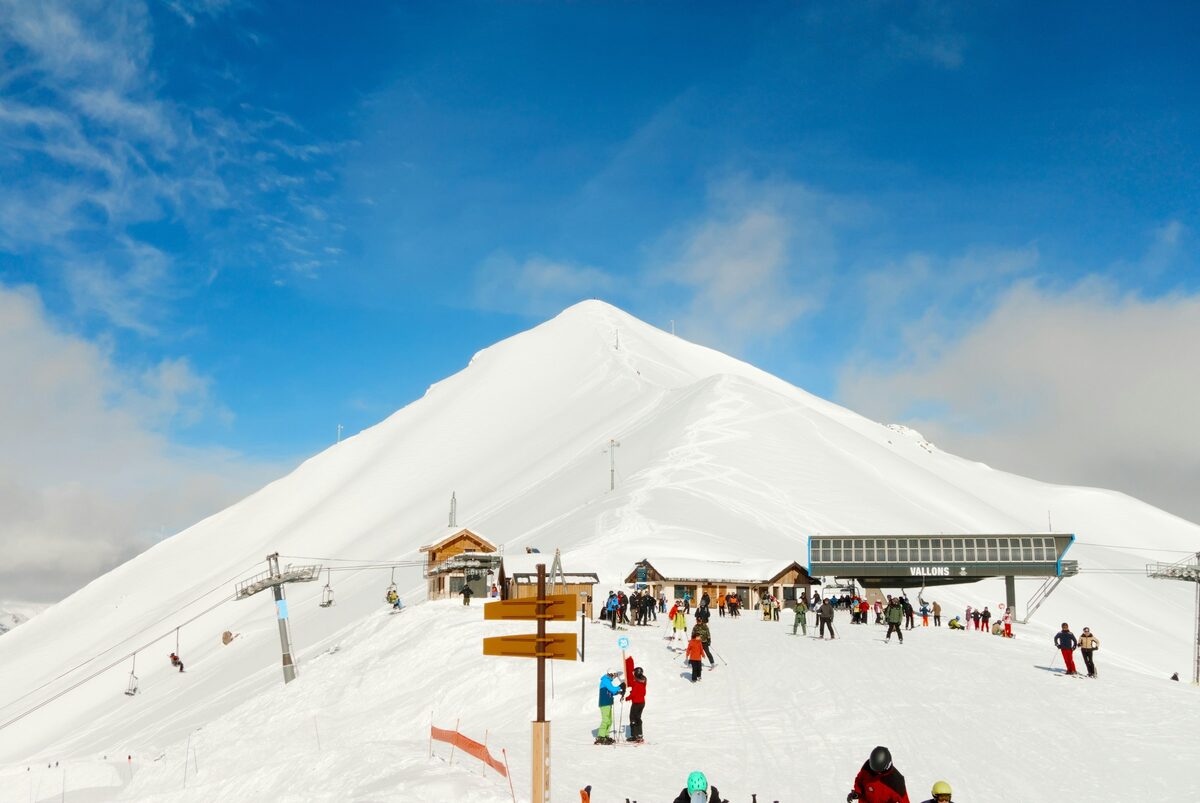 Skiers at the top of a lift below a snowy cone-shaped peak on a sunny day