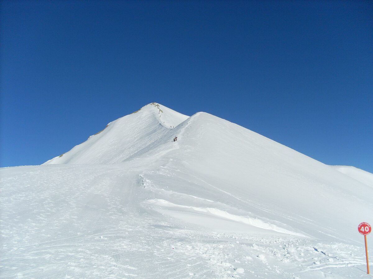 A snowy summit ridge under a deep blue sky, with a red piste marker post at the edge of the groomed snow