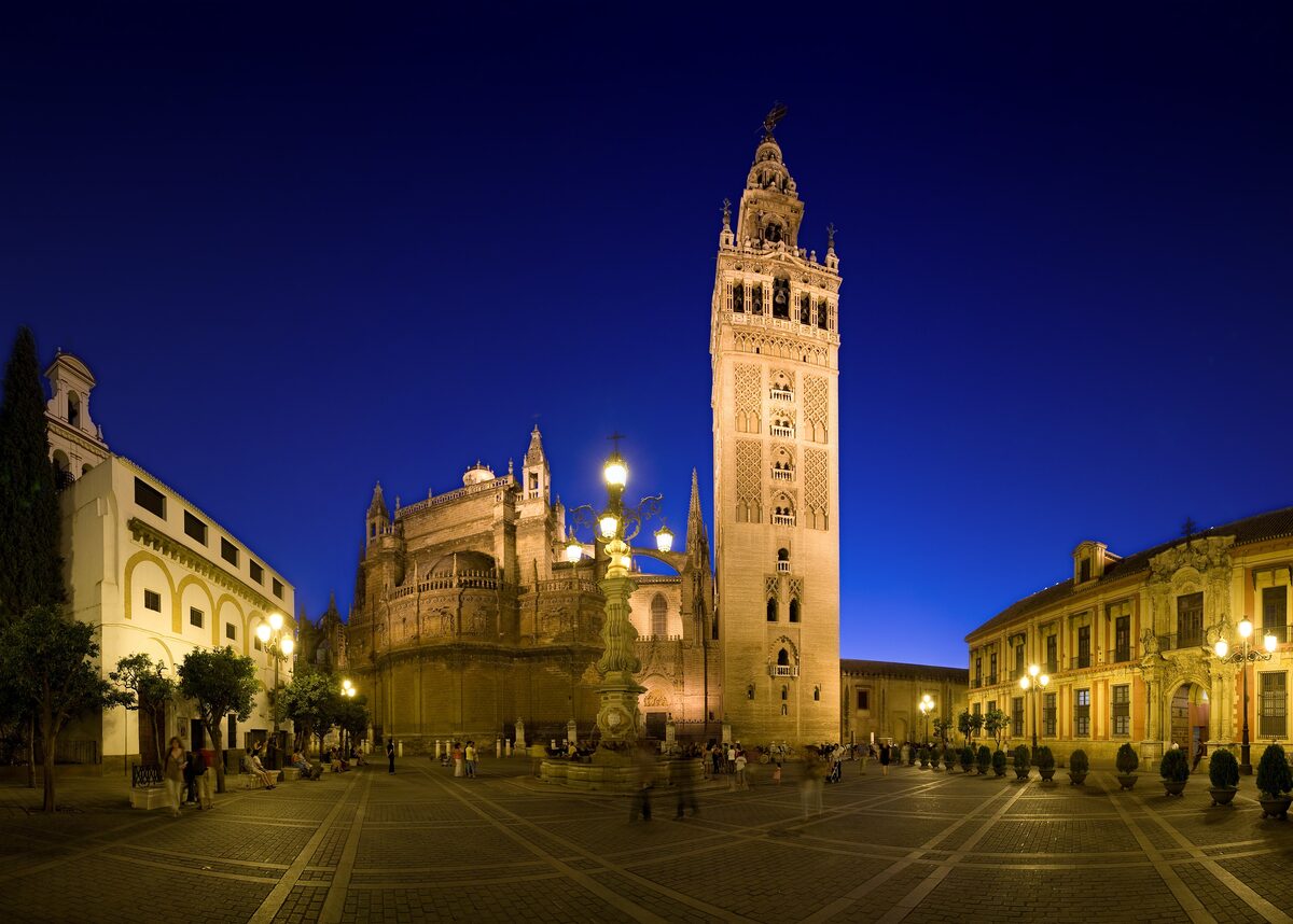 Seville Cathedral and the illuminated Giralda tower seen across Plaza Virgen de los Reyes at dusk, under a deep blue sky