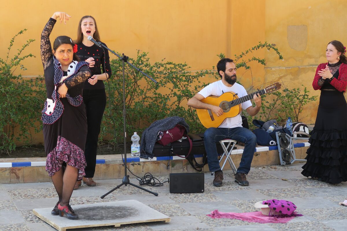A flamenco dancer in a black and pink dress performing in a Seville courtyard, with a singer and a guitarist behind her