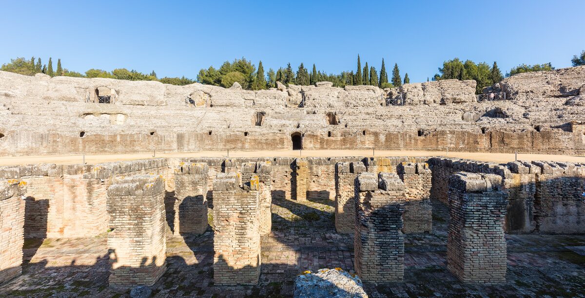 The ruined stone tiers and arches of the Roman amphitheater of Itálica under a clear blue sky, with trees above