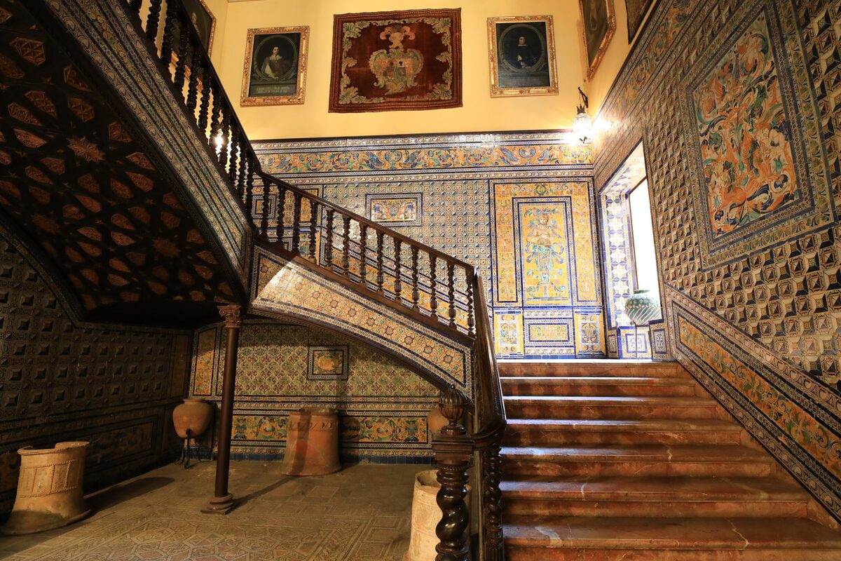 The grand staircase of the Palacio de la Condesa de Lebrija, its walls covered in colorful tiles under a carved wooden ceiling