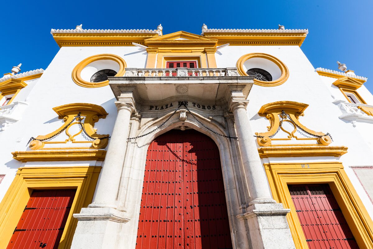 The white and ocher baroque gateway of the Maestranza bullring in Seville, with red doors under a blue sky